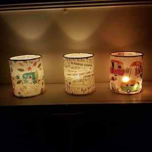 Op de afbeelding: Drie witte stoffen kaarshouders met een kampeerthema. De kaarshouders zijn aangestoken en geven een warm licht. Eén kaarshouder heeft een bloemenmotief, een andere heeft een campermotief en de derde heeft een ontwerp met de woorden "Funtive Hike Wild Glamper Camper Adventure Tree Camp Hair Don't Care Camping Nature Explore Into The Woods Explore Tent Advt Wild".