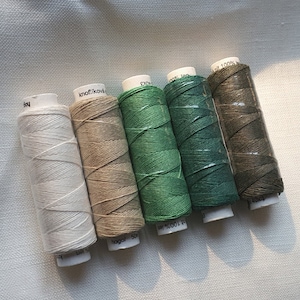 Può includere: Cinque bobine di filo in varie tonalità di verde e marrone. Le bobine sono etichettate con il nome del marchio "knorfikova" e il testo "100% len".