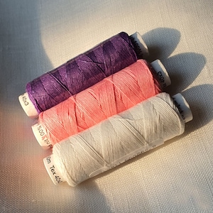 Könnte beinhalten: Drei Garnrollen, eine lila, eine rosa und eine weiße. Die Rollen sind mit dem Text "100%  40x3", "100%  40x3" und "50m Tex 40x4" beschriftet.