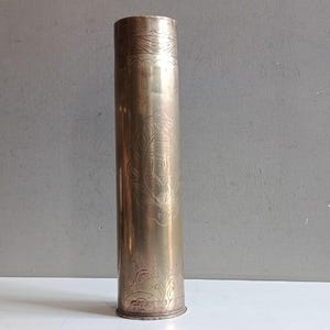 Puede incluir: Un casquillo de latón cilíndrico y alto que presenta intrincados patrones florales y de volutas grabados en los bordes superior e inferior, con un destacado grabado decorativo de forma ovalada centrado en el cuerpo metálico liso.