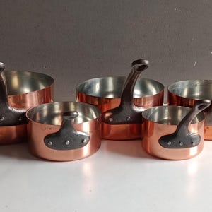 Peut inclure: Un ensemble de cinq casseroles rondes en cuivre avec des intérieurs argentés et des poignées métalliques sombres et incurvées fixées par trois rivets visibles. Les casseroles varient en taille et sont disposées sur deux rangées.