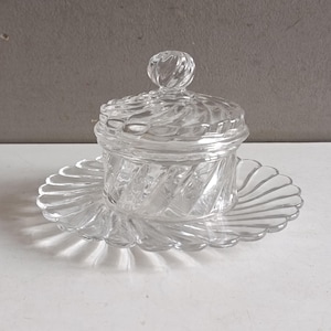 Peut inclure: Un beurrier en verre transparent avec un couvercle et une assiette assortie. Le plat et le couvercle ont un motif en spirale, et l'assiette a un bord festonné. Le plat est posé sur une surface blanche.