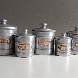 May include: A set of five cylindrical metal canisters with lids, each featuring a decorative embossed band and a brass-toned label plate. The labels are embossed with the French words SUCRE, THE, CAFE, EPICES, and FARINE in capital letters.
