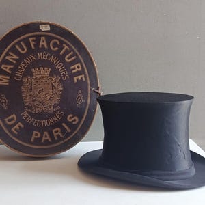 May include: A black silk top hat positioned next to a round, dark brown cardboard hat box. The box features gold text reading MANUFACTURE DE PARIS CHAPEAUX MECANIQUES PERFECTIONNES and a central crest with a ship and crown illustration.