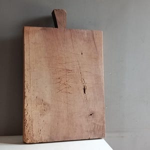 Peut inclure: Une planche à découper rectangulaire en bois avec une poignée. La planche est de couleur marron clair avec des veines et des rayures visibles. La poignée est une petite pièce de bois rectangulaire.