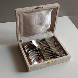 Può includere: Un set di cucchiaini d'argento in una scatola beige con fodera in raso. I cucchiaini sono disposti in file su una base di feltro verde. La scatola è aperta, rivelando i cucchiaini e il logo Christofle.