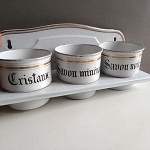 May include: A white enamel wall-mounted shelf with three round containers. Each container has a gold band and black text: "Cristaux", "Savon minéral", and "Savon noir". The shelf has a decorative gold trim.