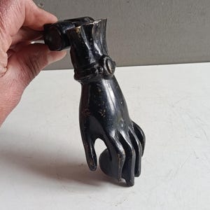 Puede incluir: Un aldaba de puerta negra de estilo antiguo con forma de mano. La aldaba es de metal y tiene un diseño detallado, con nudillos visibles y un anillo en la muñeca. El acabado tiene un aspecto ligeramente envejecido.