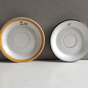 Peut inclure: Deux soucoupes en céramique blanche avec des centres circulaires. La soucoupe de gauche présente un bord jaune et le texte noir 2.50. La soucoupe de droite a un bord noir et le texte noir 4f. Les deux ont un fin cercle doré au centre.