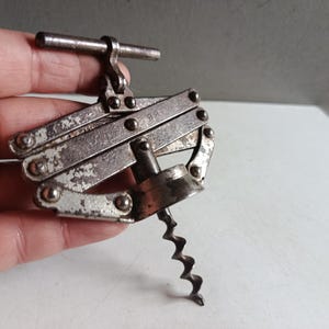 May include: A vintage silver-coloured corkscrew with a lever design. The corkscrew has a worm-style bit and a handle. The metal shows signs of wear, giving it a rustic appearance. It is designed to remove corks from wine bottles.