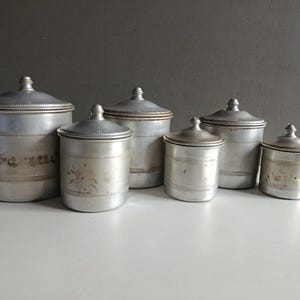 May include: A set of six vintage metal canisters with lids. The canisters are silver-toned with decorative etched bands and floral patterns. They vary in size, arranged from largest to smallest, and sit on a white surface.