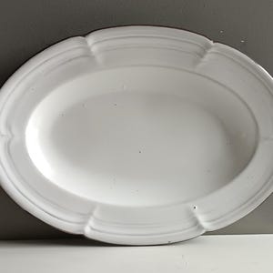 Puede incluir: Plato ovalado blanco para servir con borde festoneado. El plato tiene un borde ligeramente elevado y una superficie lisa y brillante. El borde presenta un patrón decorativo y repetitivo. El plato es probablemente de cerámica o porcelana.