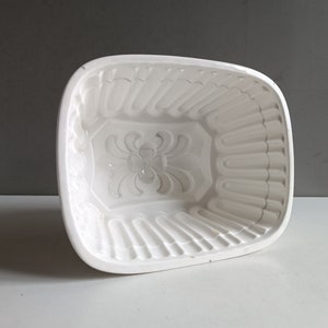 May include: White ceramic mold with a decorative floral design. The mold is shaped like a rectangle with rounded corners and has a fluted edge.