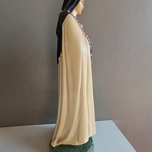 May include: A standing figurine of a figure wearing a long, cream-colored robe and a black head covering. The figure has a pink beaded rosary draped around the neck and shoulders, and stands on a textured, dark green hexagonal base.
