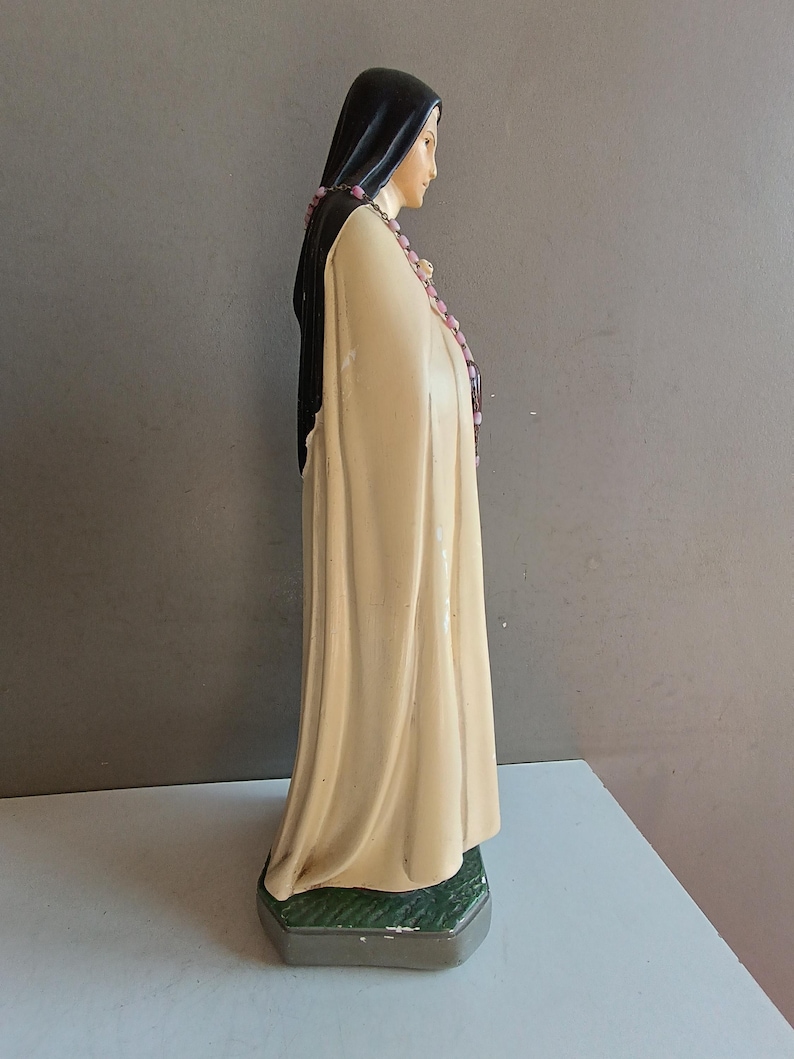 May include: A standing figurine of a figure wearing a long, cream-colored robe and a black head covering. The figure has a pink beaded rosary draped around the neck and shoulders, and stands on a textured, dark green hexagonal base.