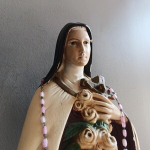 May include: A religious statue of a figure wearing a black habit and a cream-colored cloak, holding a cluster of sculpted roses and a crucifix against the chest. A rosary with light pink beads is draped over the shoulders of the statue.