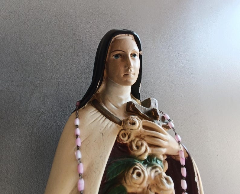 May include: A religious statue of a figure wearing a black habit and a cream-colored cloak, holding a cluster of sculpted roses and a crucifix against the chest. A rosary with light pink beads is draped over the shoulders of the statue.