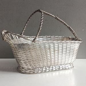 May include: A silver-toned metal basket with a woven-style pattern and a rectangular base. The basket features a high, arched handle with a twisted rope design and an elevated, open-ended spout on one side, creating a distinct elongated shape.