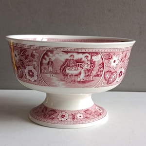 Peut inclure: Un bol à pied blanc et rouge avec un décor floral et pastoral. Le bol présente un large bord et un pied en forme de piédestal. Le motif rouge comprend des fleurs et une scène de personnes à table. Le bol est probablement en céramique.