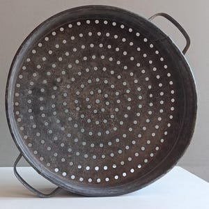 Pode incluir: Um escorredor redondo de metal cinza-escuro com uma bacia larga e rasa, apresentando um padrão uniforme de orifícios de drenagem circulares em toda a base. O escorredor inclui duas alças de metal curvas em lados opostos da borda.