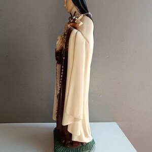 May include: A religious figurine depicting a figure in a black habit and cream-colored cloak, holding a crucifix and a bouquet of flowers. A pink beaded rosary hangs from the hands, and the figure stands on a textured green base.