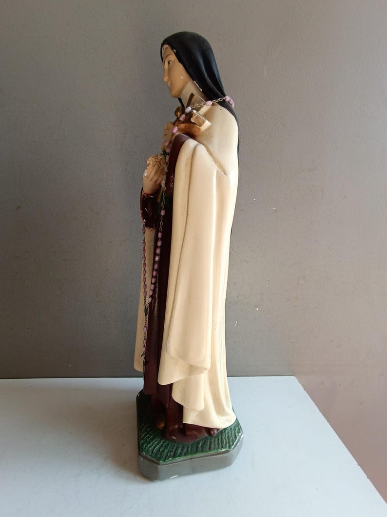 May include: A religious figurine depicting a figure in a black habit and cream-colored cloak, holding a crucifix and a bouquet of flowers. A pink beaded rosary hangs from the hands, and the figure stands on a textured green base.