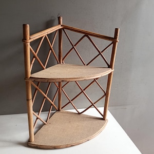 Peut inclure: Étagère d'angle vintage en bambou naturel. L'étagère comporte deux étagères semi-circulaires et un motif en treillis de losanges. Le bambou est de couleur brun clair et l'étagère est conçue pour s'adapter à un coin. Elle a un crochet pour suspendre.