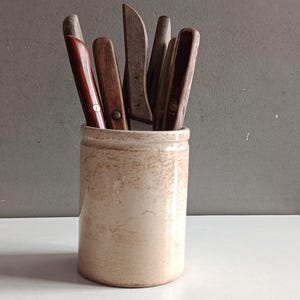 Peut inclure: Un pot en céramique cylindrique avec une finition beige et brune tachetée, rempli d'une collection de couteaux de cuisine dotés de divers manches en bois dans des tons de brun clair, moyen et foncé, chacun fixé par des rivets métalliques.