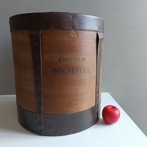 Puede incluir: Un contenedor de almacenamiento de madera vintage con bandas metálicas y el texto "DOUBLE DECALITRE" impreso en el lateral. El contenedor es marrón y tiene una manzana roja a su lado.