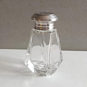 Pode incluir: Uma caneca de cerâmica branca com uma alça preta e o texto "But First, Coffee" em preto, com uma fonte manuscrita. A caneca está sobre uma superfície branca.