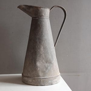 Può includere: Una tazza di ceramica bianca con manico nero e la scritta "But First, Coffee" in grassetto nero. La tazza è piena di un liquido scuro, probabilmente caffè, e poggia su una superficie chiara.