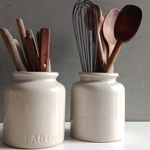 Pode incluir: Dois potes de cerâmica na cor creme contendo utensílios de cozinha. O pote esquerdo apresenta o texto em relevo LAB Lag e contém ferramentas de madeira. O pote direito contém um batedor de metal e várias colheres de madeira.