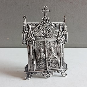 Puede incluir: Un tríptico de metal en tono plateado con intrincados detalles arquitectónicos góticos, que incluyen arcos apuntados y agujas. Los paneles frontales muestran retratos en relieve del Sagrado Corazón de Jesús y el Inmaculado Corazón de María.