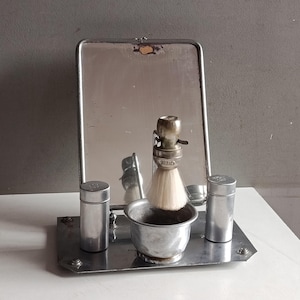Op de afbeelding: Vintage scheerset met een spiegel, borstel, kom en twee containers. De set is gemaakt van zilverkleurig metaal en staat op een rechthoekige tray. De spiegel is rechthoekig met een scharnierende standaard. De scheerkwast heeft een witte haren kop.
