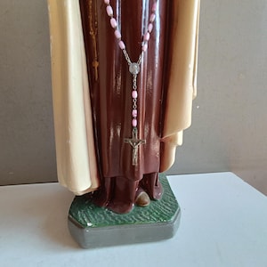 May include: A religious statue featuring a figure in a dark brown robe and a cream-colored cloak. A rosary with light pink beads and a silver-toned crucifix hangs around the neck. The figure stands on a textured green base with a gray border.