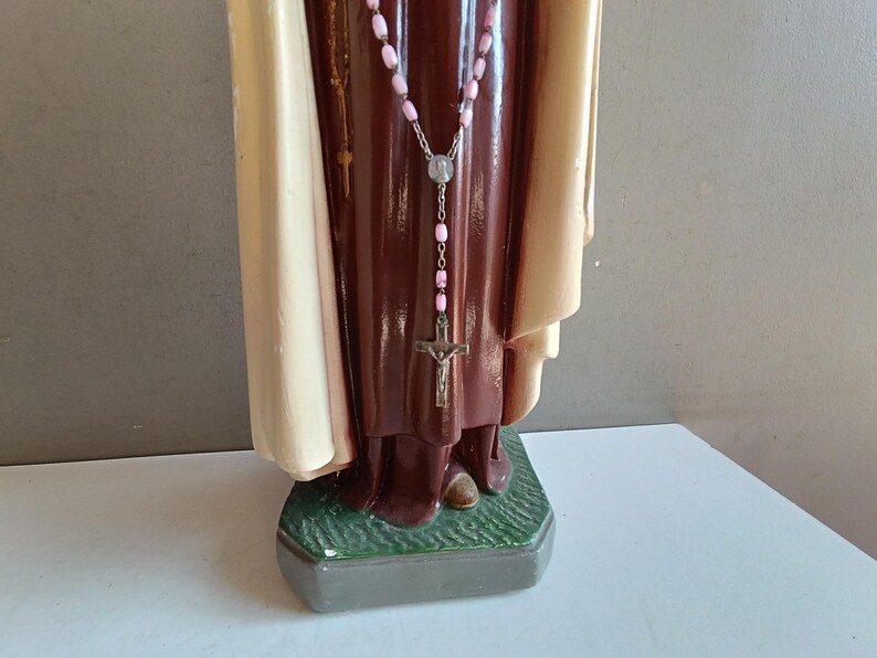 May include: A religious statue featuring a figure in a dark brown robe and a cream-colored cloak. A rosary with light pink beads and a silver-toned crucifix hangs around the neck. The figure stands on a textured green base with a gray border.
