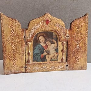 Puede incluir: Un tríptico de madera vintage con una imagen religiosa de una mujer y un niño. El marco dorado presenta intrincados diseños florales y un acento rojo. El panel central muestra las figuras en una pintura detallada y colorida.