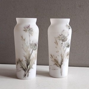 Op de afbeelding: Twee witte glazen vazen met een matte afwerking. Elke vaas heeft een handgeschilderd bloemenontwerp in grijstinten en bruin, met een distelachtige bloem en bladeren. De vazen zijn cilindrisch met een licht uitlopende bovenkant.