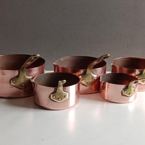 Op de afbeelding: Een set van vijf koperen steelpannen met messing handvatten. De pannen variëren in grootte, met een gepolijste koperen buitenkant en messing handvatten die met klinknagels zijn bevestigd. De binnenkant van de pannen is donkerder koperkleurig.