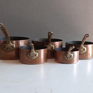 Può includere: Un set di quattro pentole in rame graduate con manici in ottone. Ogni pentola presenta un corpo cilindrico con un esterno in rame lucido e un robusto manico in ottone fissato con tre rivetti visibili sul lato dell'utensile da cucina.