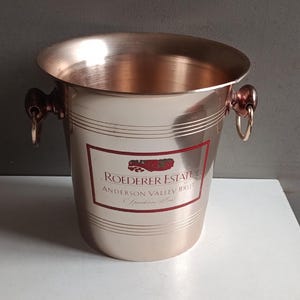 Może przedstawiać: Miedziany wiaderko na wino z etykietą z napisem "Roederer Estate Anderson Valley Brut Sparkling Wine". Wiaderko ma dwa uchwyty i polerowane wykończenie. Wiaderko stoi na białej powierzchni.
