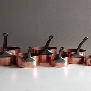 May include: A set of six copper saucepans with dark metal handles. The pans are arranged in a row, varying in size from large to small. The copper has a warm, reddish-brown hue, and the handles are riveted to the sides.