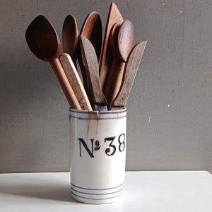 Pode incluir: Um suporte cilíndrico de cerâmica branca para utensílios com listras horizontais azuis e o texto preto "N°38" impresso na frente. O suporte está cheio de uma coleção de vários utensílios de cozinha de madeira, incluindo colheres, uma espátula e facas.