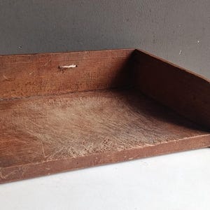 Pode incluir: Uma bandeja de escritório retangular de madeira com acabamento em marrom escuro e uma superfície desgastada e arranhada. A bandeja possui um painel traseiro alto com um pequeno orifício central e dois painéis laterais que se inclinam em direção à borda frontal aberta.