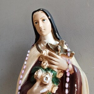 May include: A religious figurine of a figure wearing a black veil and a cream-colored mantle over a dark brown garment. The figure holds a bouquet of white roses and a brown wooden crucifix, with a string of light pink beads draped over the shoulders.