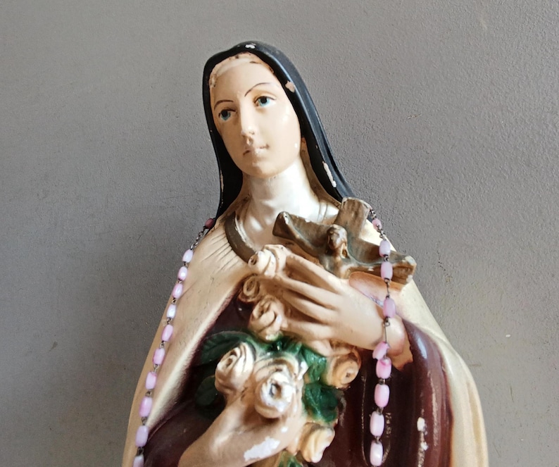 May include: A religious figurine of a figure wearing a black veil and a cream-colored mantle over a dark brown garment. The figure holds a bouquet of white roses and a brown wooden crucifix, with a string of light pink beads draped over the shoulders.