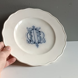 Op de afbeelding: Een wit keramiek bord met een blauw en wit monogram in het midden. Het bord heeft een gegolfde rand.