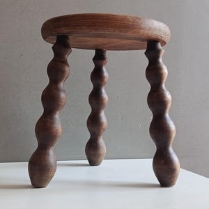 May include: A small, round wooden stool featuring a smooth, circular top and three distinct legs. Each leg is crafted with a series of stacked, rounded, bulbous shapes that taper toward the base, finished in a dark, natural wood grain texture.