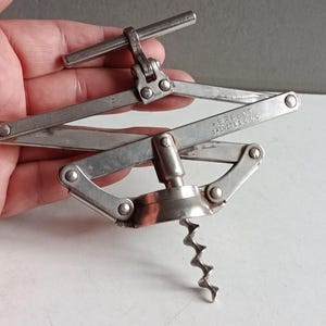 Könnte beinhalten: Ein silberfarbener Metallkorkenzieher mit einem zusammenklappbaren Scherenmechanismus. Das Werkzeug verfügt über eine zentrale Spiralspindel und einen oberen Griff. Der Metallarm ist deutlich mit dem Text PERFECT BREVETE S.G.D.G. geprägt.