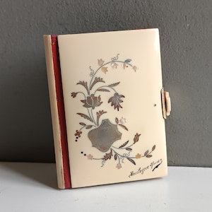 Può includere: Una piccola scatola color avorio con una spina dorsale rossa e un motivo floreale. La scatola ha una chiusura argentata e le parole "Souvenirs d'Yverdon" scritte sul davanti.